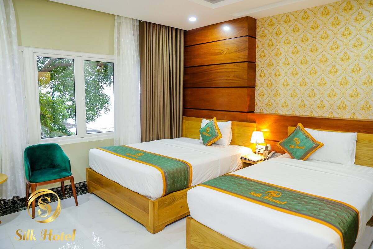 A hotel room with two beds, a green chair, and a window with curtains.