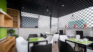 An outdoor café with wicker chairs and tables, a wooden bench, and potted plants. - Silk Hotel (Ho Chi Minh City)