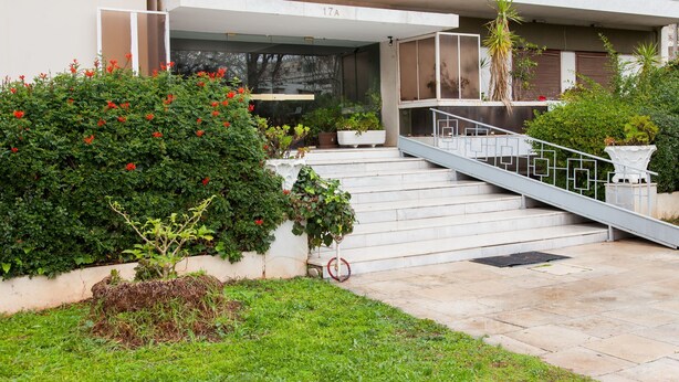 A modern building entrance with a staircase leading to a balcony, surrounded by greenery and potted plants. - Glyfada Square Modern And Cozy Apartment (Glyfada)