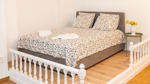 A bedroom with a patterned bedspread, a white railing, a small bedside table with a vase of flowers, and a grey sofa in the background. - Glyfada Square Modern And Cozy Apartment (Glyfada)