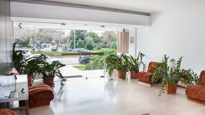 A modern interior with a large glass door leading to a parking area, several potted plants, and a seating area with brown upholstered chairs. - Glyfada Square Modern And Cozy Apartment (Glyfada)