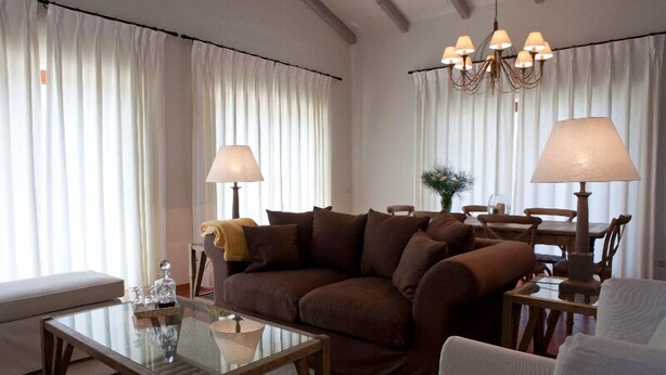 Una sala de estar con un sofá marrón, una mesa de centro de cristal, un sillón blanco y una mesita de noche de madera con una lámpara.