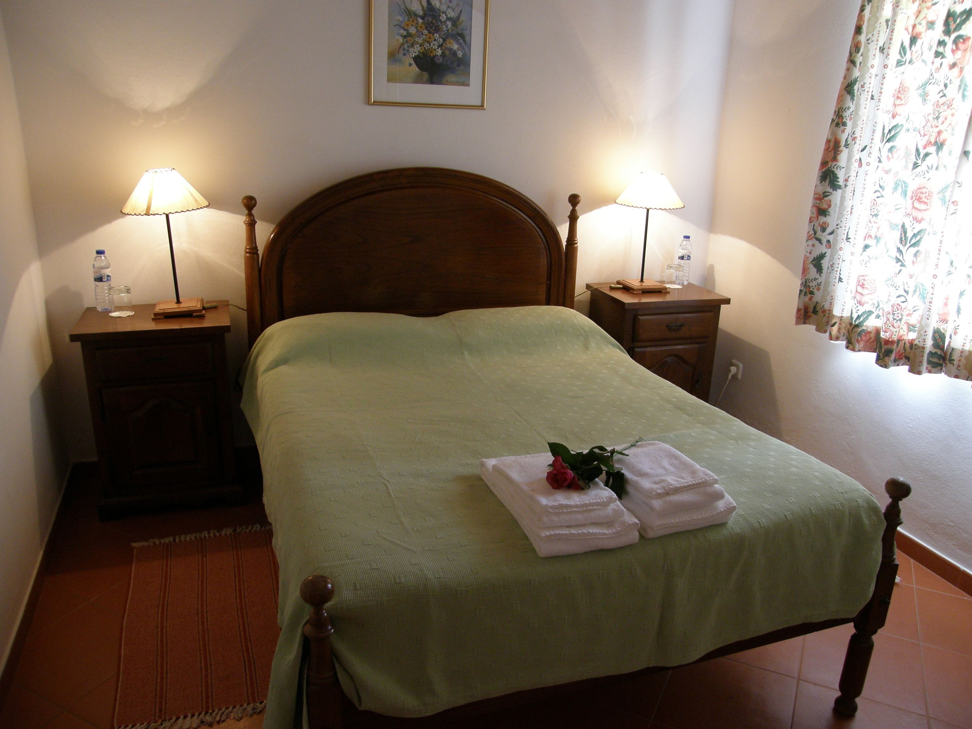 A neatly made bed with a green bedspread, two bedside tables with lamps, a wooden headboard, and a floral curtain.