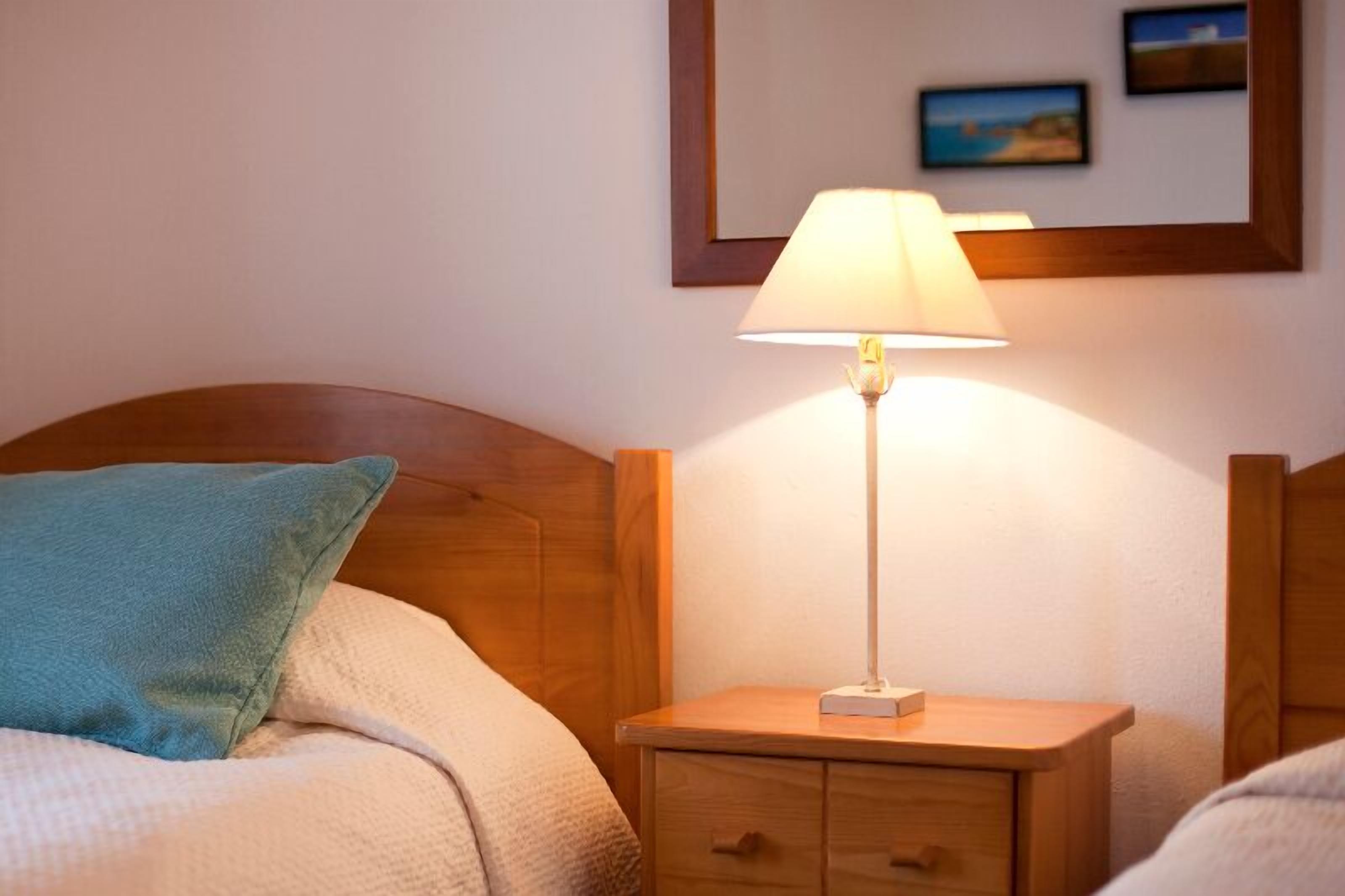 Una cama bien tendida con un cojín azul, una mesita de noche con una lámpara y un cuadro colgado en la pared.