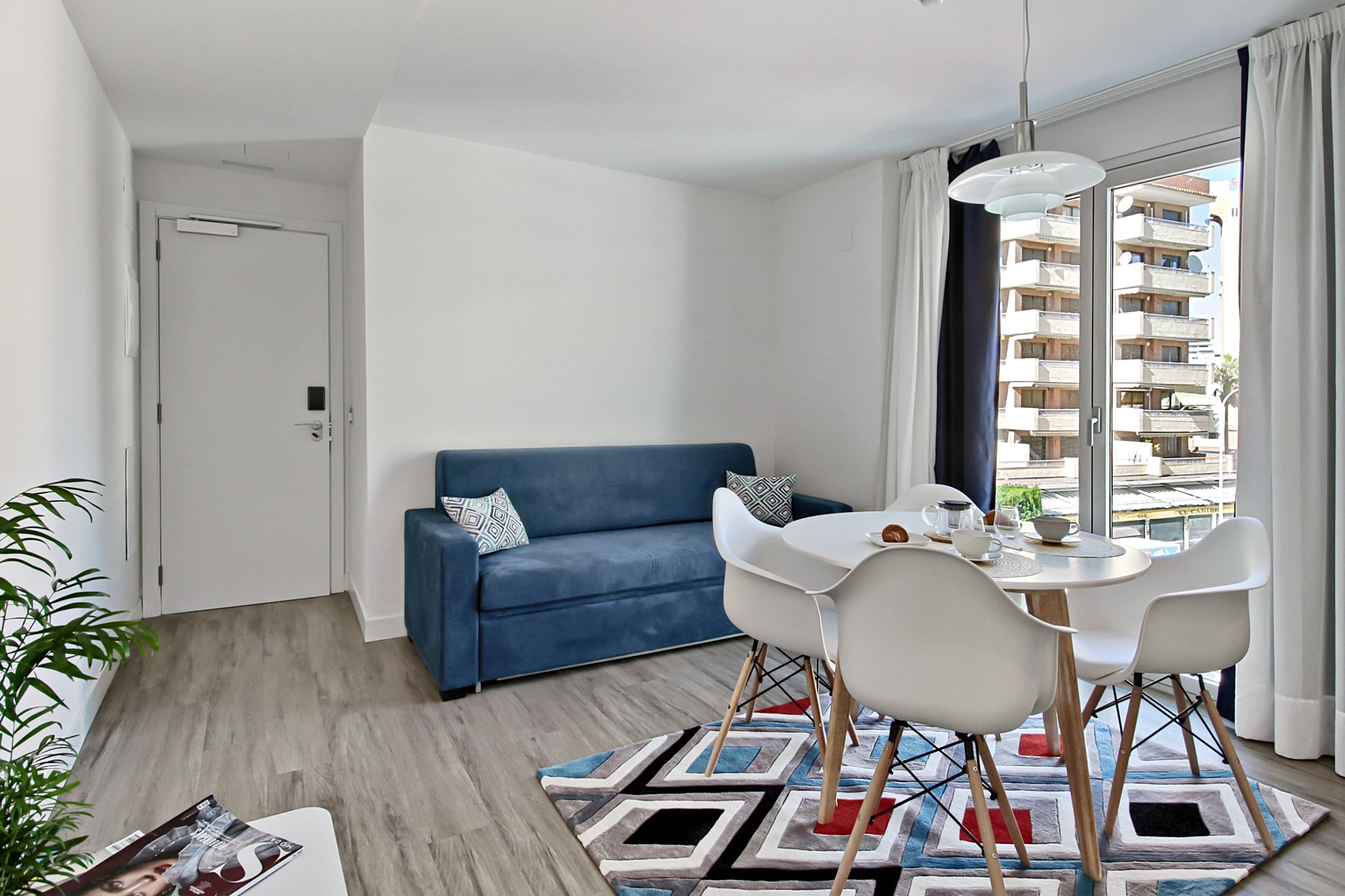 Ein modernes Wohnzimmer mit einem blauen Sofa, weißen Sesseln, einem runden Tisch und einem gemusterten Teppich.