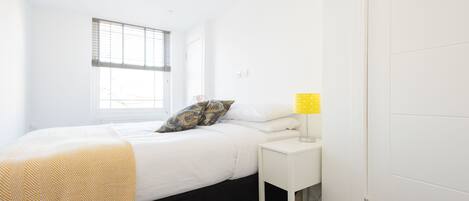 A bedroom with a bed, a bedside table with a lamp, a window with blinds, and a door.