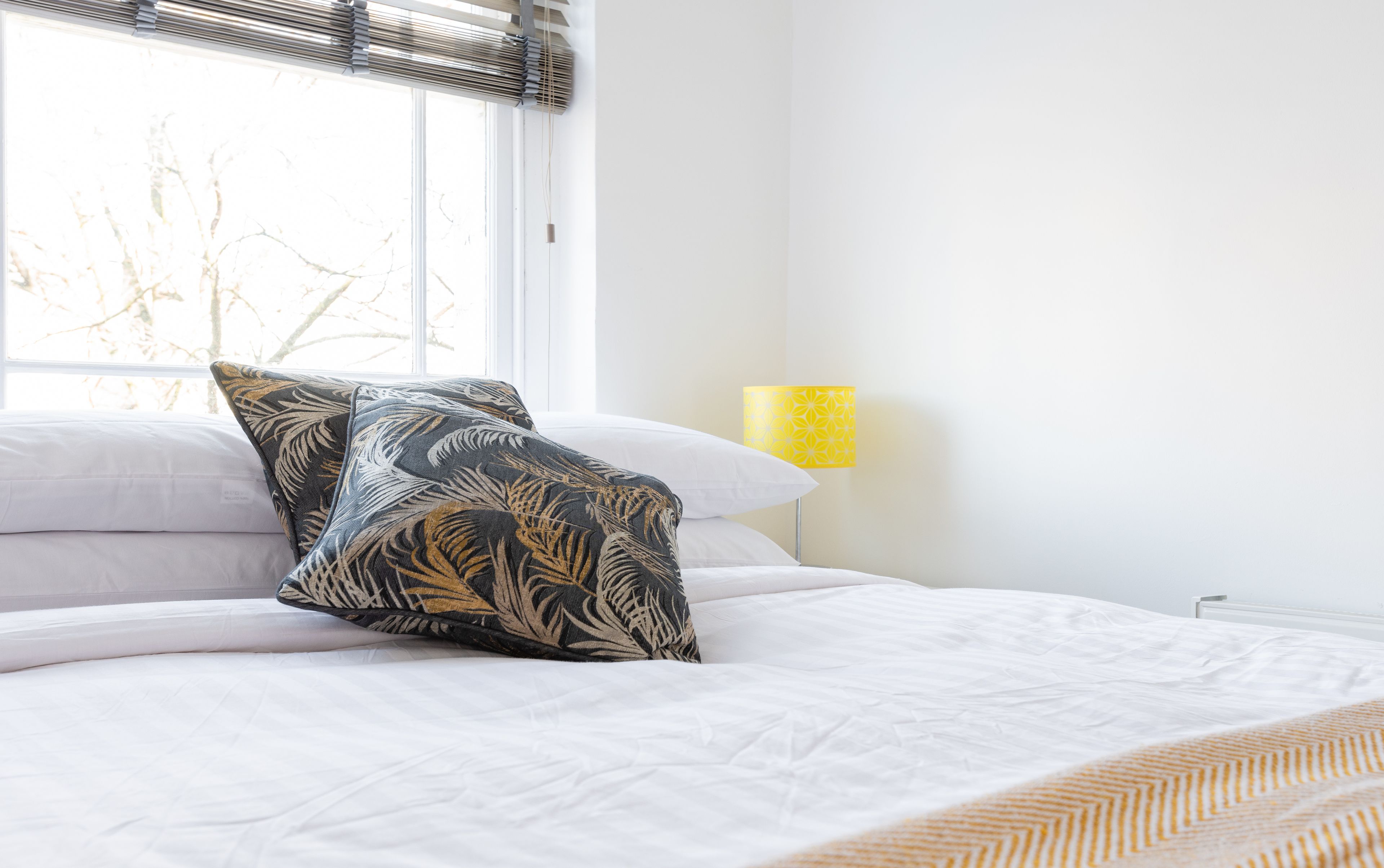 Een bed met wit beddengoed en twee decoratieve kussens, een gele lamp en een raam met jaloezieën.
