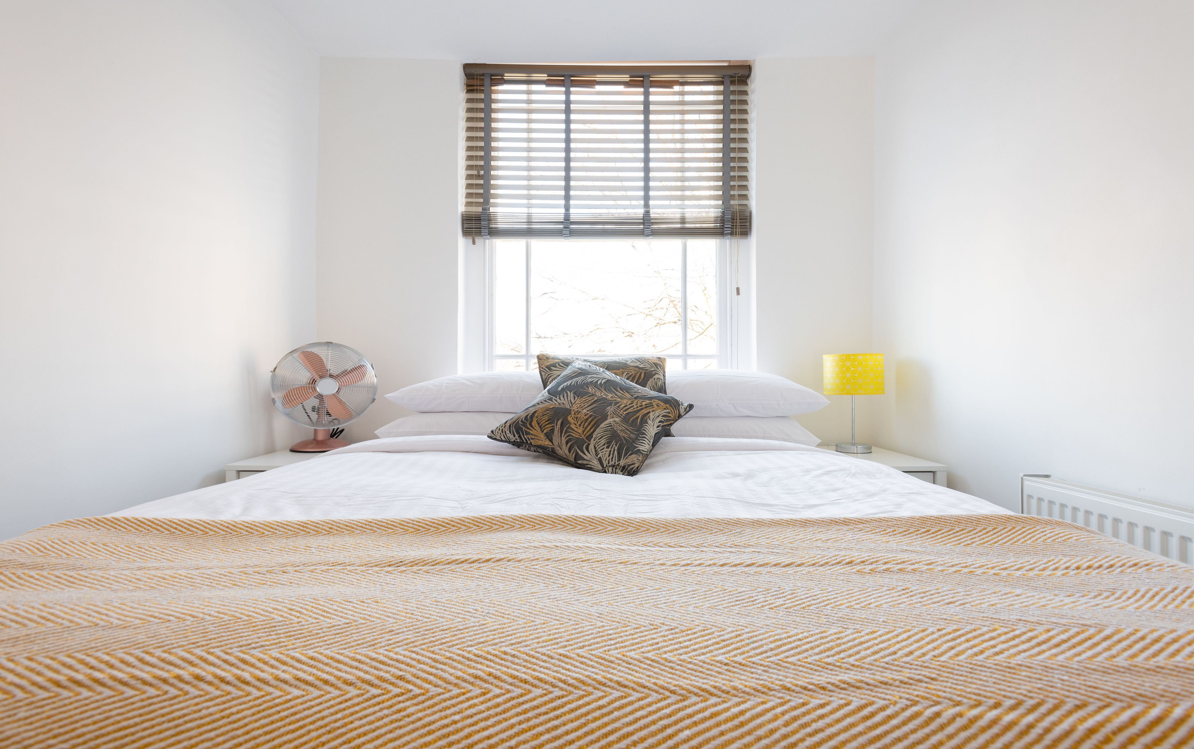 A bedroom with a bed, a standing fan, a window with blinds, and a bedside lamp.