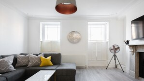 A modern living room with a grey sectional sofa, a small white table, and a standing fan. - The Marylebone Residence (London)