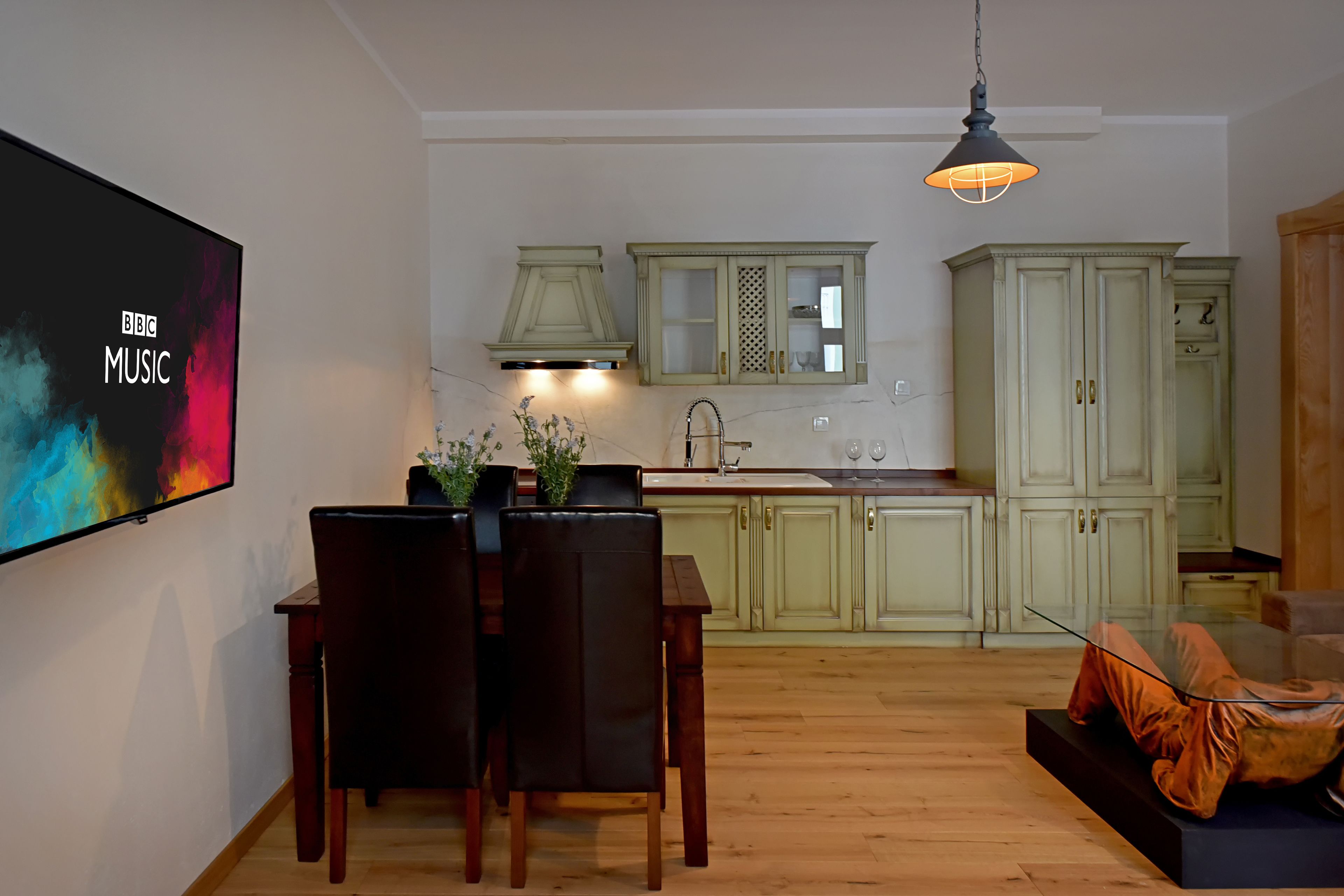 A modern kitchen with green cabinets, a dining area with a TV displaying 'BBC MUSIC', and a cozy seating area.