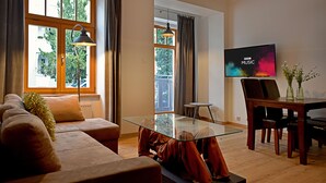Una sala de estar moderna con una mesa de centro de vidrio, un sofá, una mesa de comedor con sillas, un televisor de pantalla plana y ventanas grandes con cortinas.