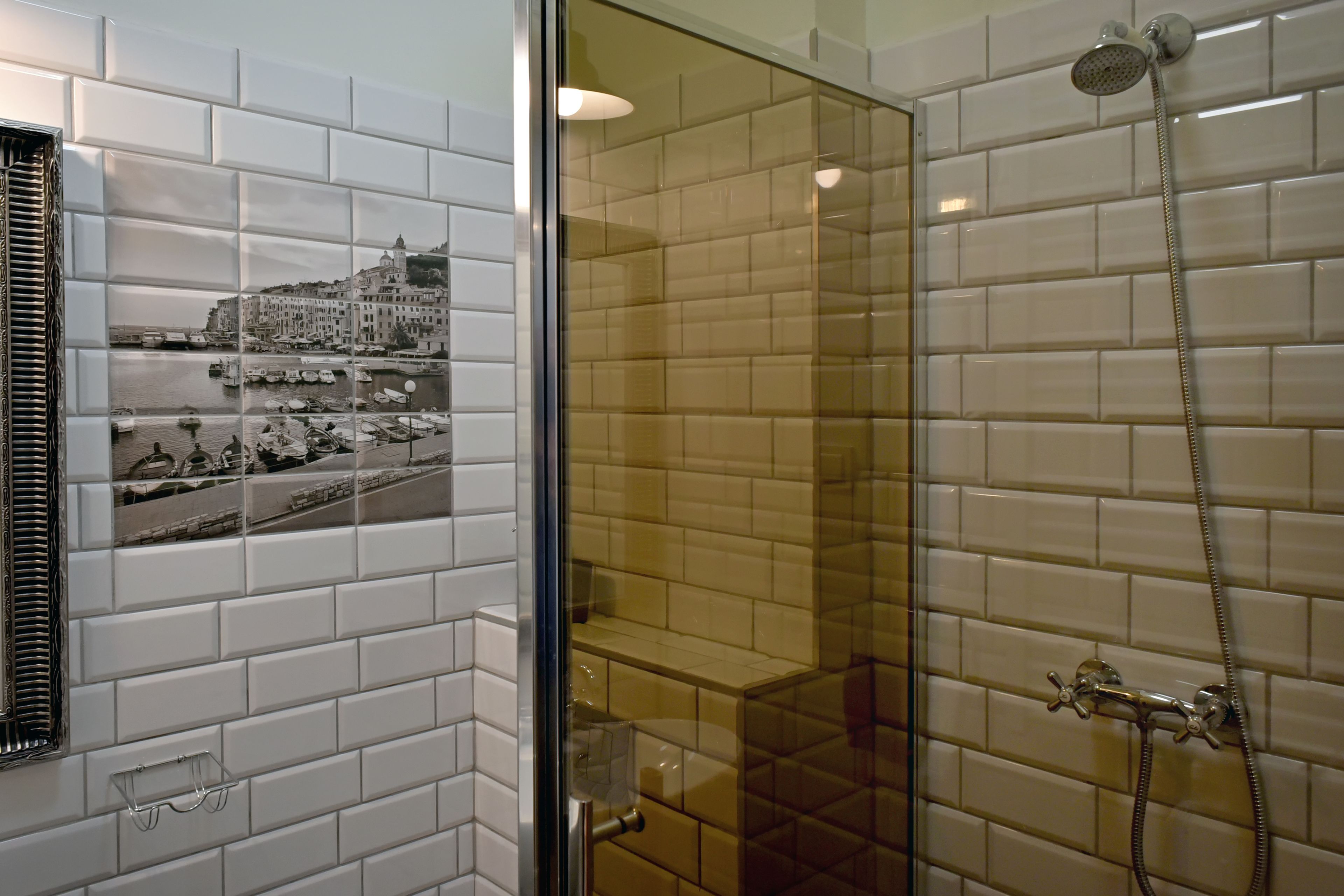 Un baño con azulejos blancos tipo metro, cerramiento de ducha de vidrio y una fotografía enmarcada de un pueblo costero.