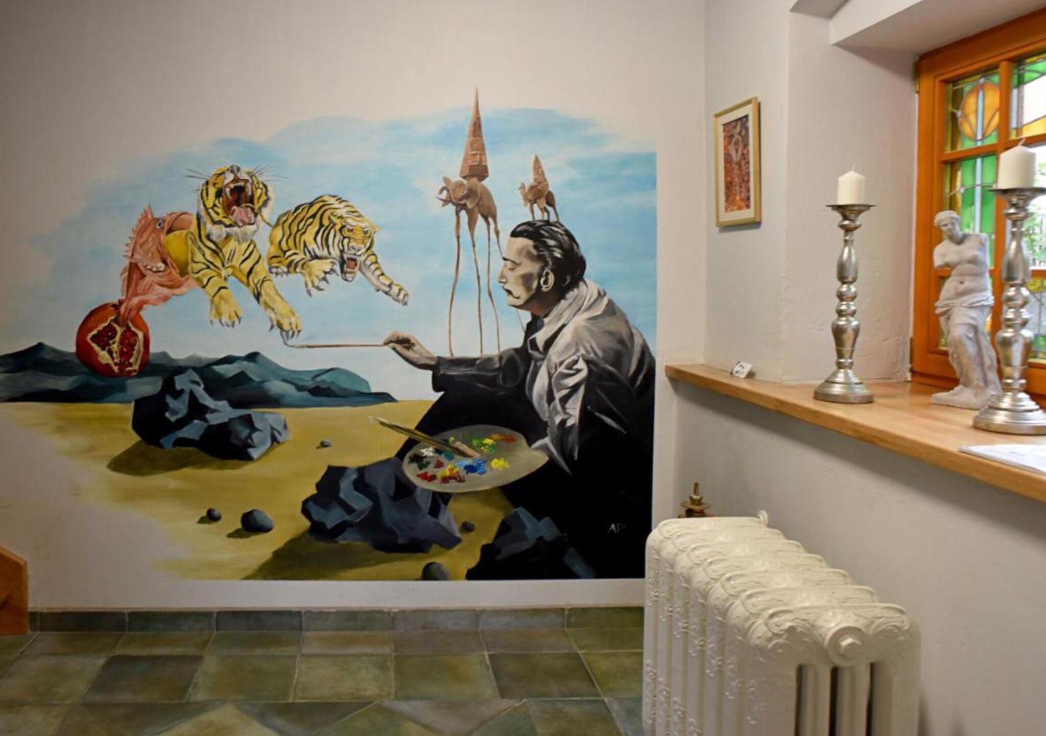 Un mural con un pintor, un tigre y una criatura roja. Se observa un radiador, una ventana que da a una vista verde y una estatua sobre una estantería.