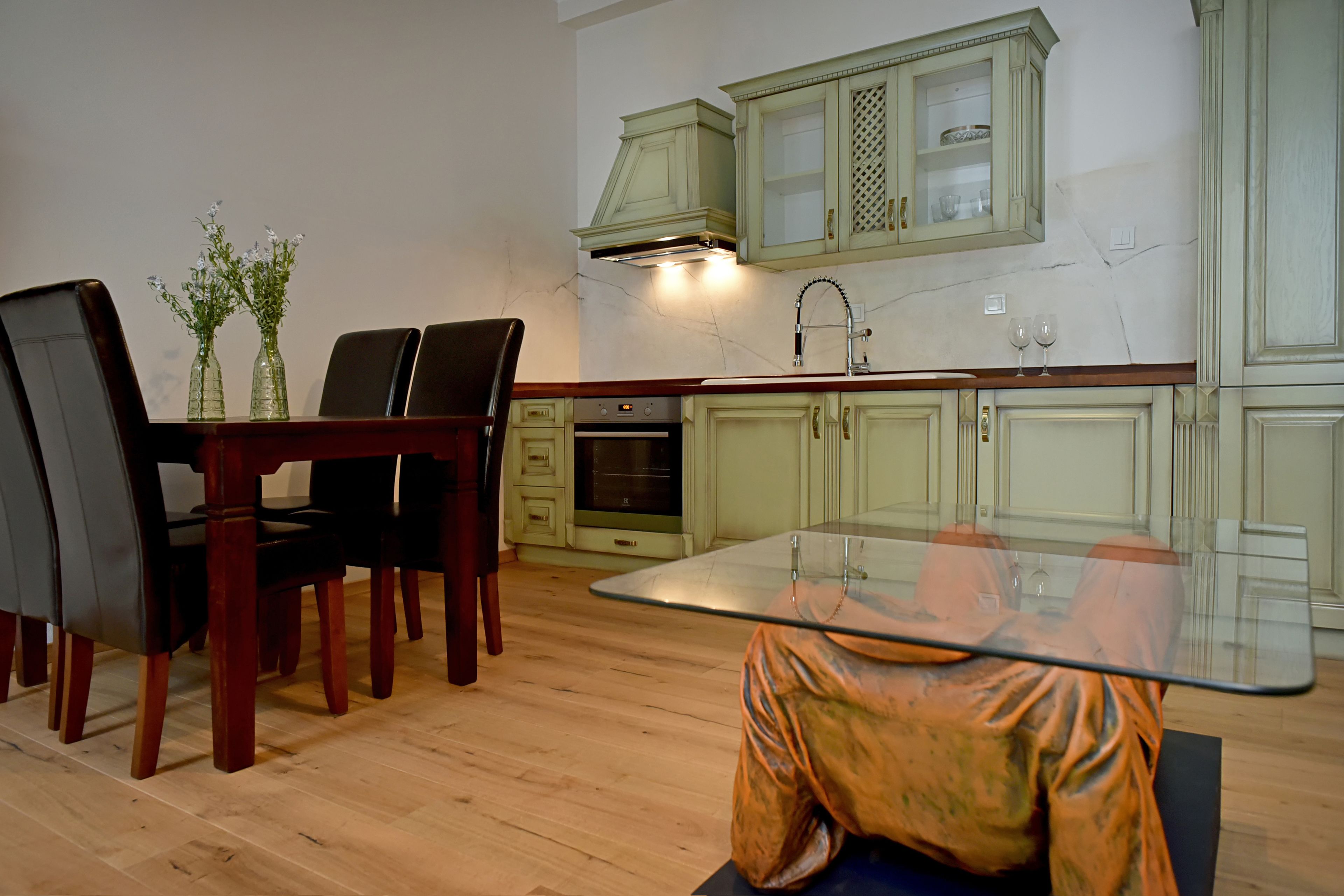 A dining area with a glass table, chairs, and a vase with flowers. There is a kitchen with cabinets and a stove in the background.