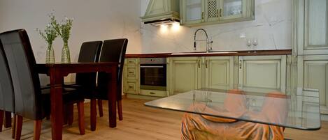 A dining area with a glass table, chairs, and a vase with flowers. There is a kitchen with cabinets and a stove in the background.