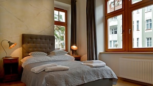Una cama bien tendida con un edredón gris estampado, dos lámparas de noche y vistas de un edificio a través de una ventana.