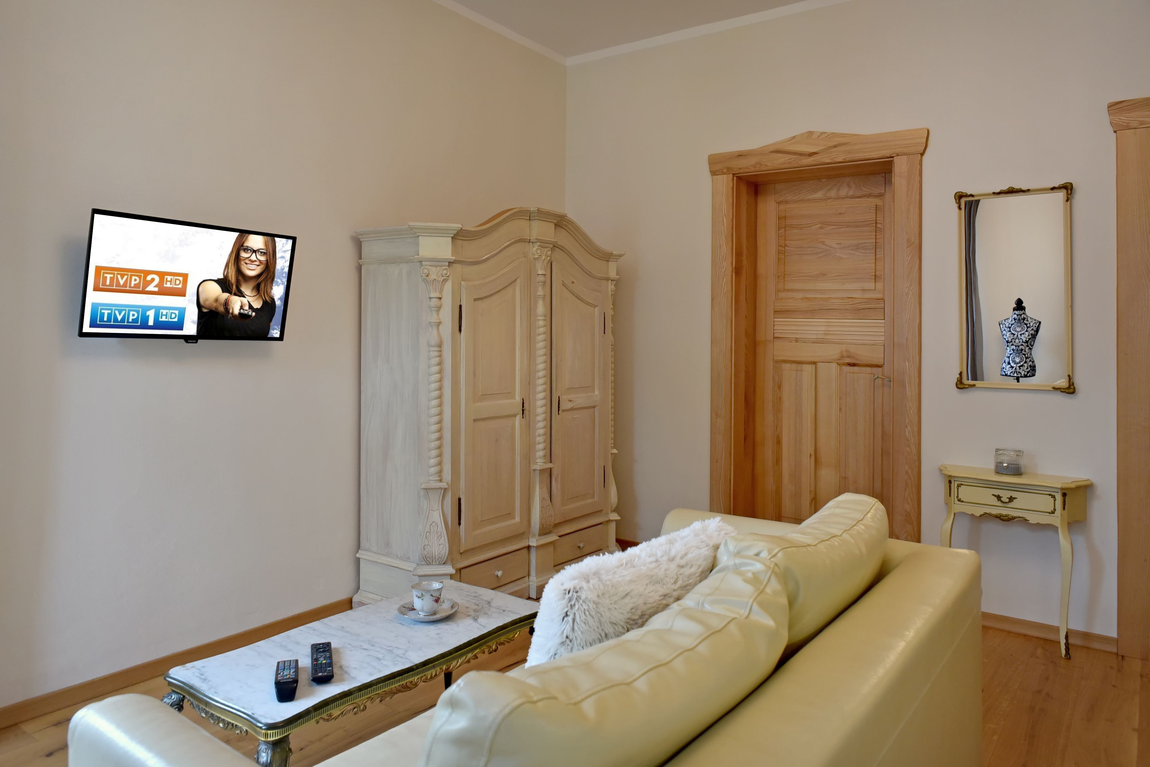 A living room with a TV displaying TV channels, a white sofa, a wooden wardrobe, a wooden door, and a small table with a vase.