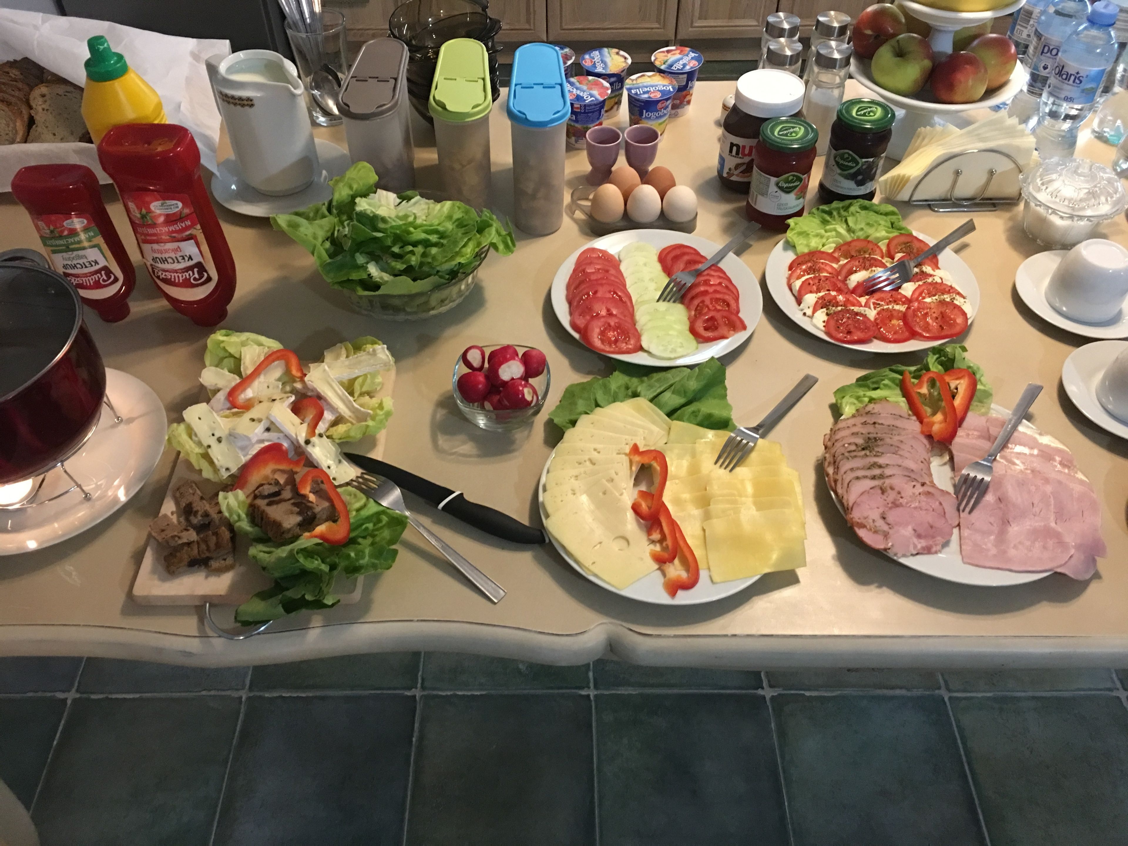 A kitchen counter with various food items and condiments, including plates of sliced meat, cheese, and vegetables, as well as bottles of ketchup and mustard.