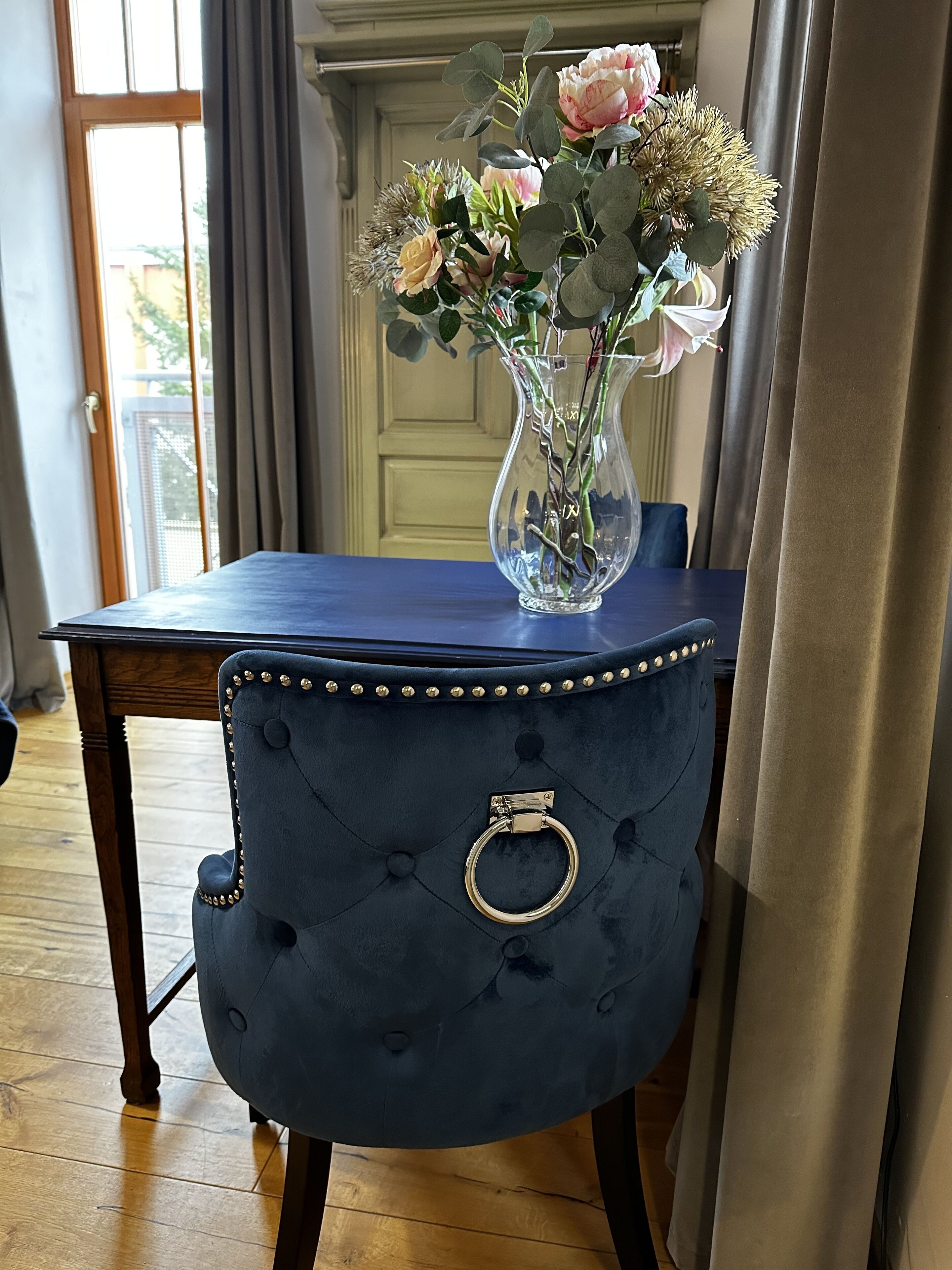 Un fauteuil bleu foncé recouvert de tissu, orné d’un anneau doré, un vase transparent rempli de fleurs et une table bleue.