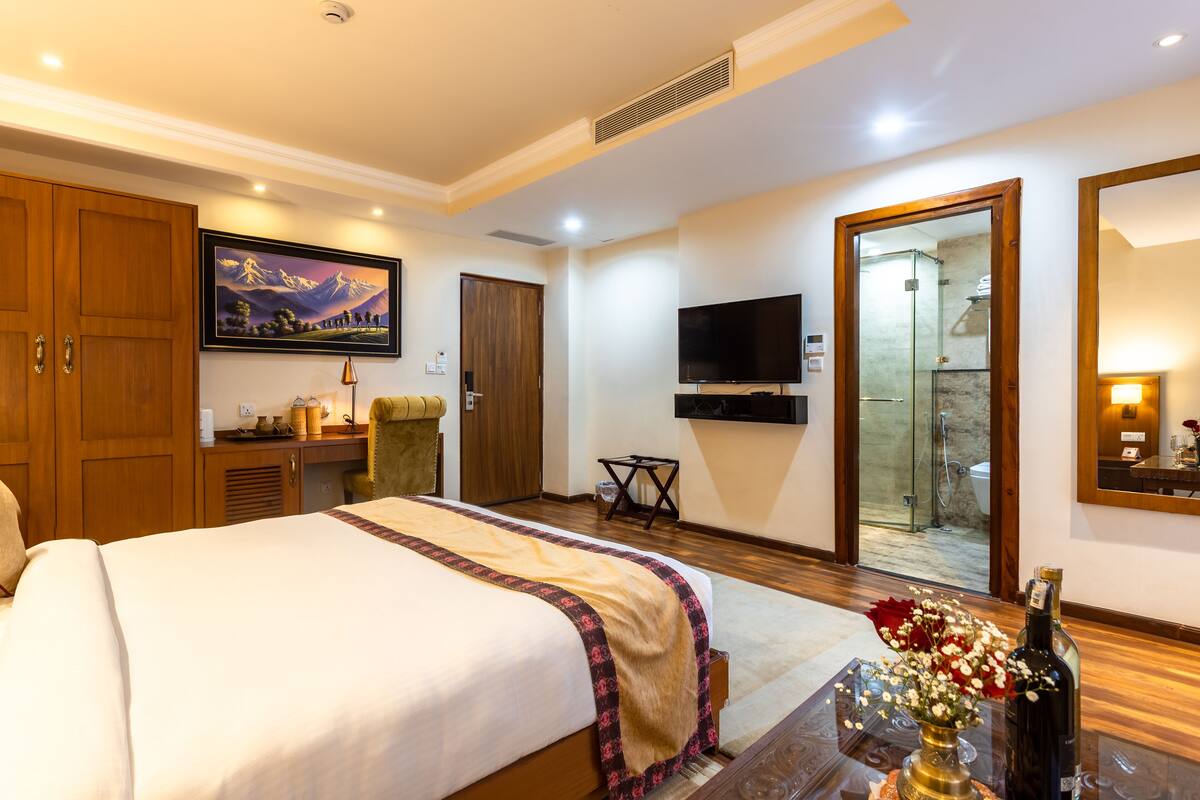 A hotel room with a large bed, a flat-screen TV, a wooden wardrobe, a bathroom with a glass shower, and a framed picture on the wall.