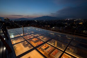 Terraço no telhado com piso de vidro, áreas de estar mobiliadas e vista para a cidade ao anoitecer.