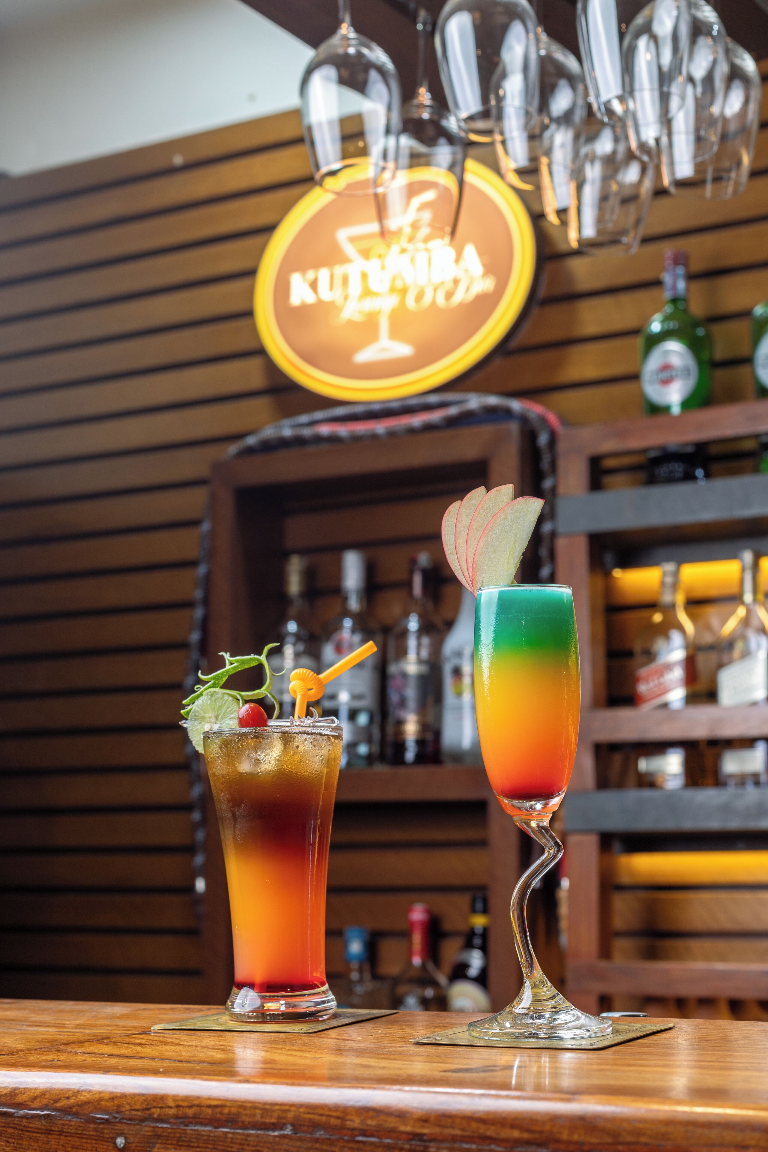 Deux cocktails sur un comptoir de bar, avec un fond en bois et une enseigne au néon en arrière-plan.