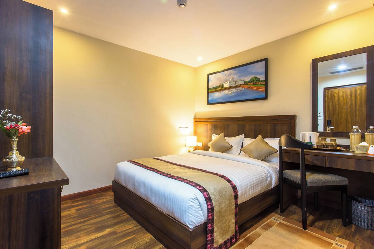 A hotel room with a large bed, a desk with a chair, a vase with flowers, and a painting on the wall.