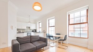 A modern living room with a grey sofa, a wooden coffee table, and a dining area with a glass table and chairs. There is a kitchen in the background with white cabinets and a hanging light fixture. - The Enya Suite - hiphipstay (Dublin)