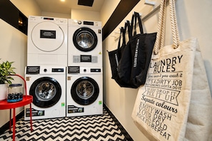 A laundry area with two washing machines, a red chair, a plant, and a large tote bag with laundry rules. - The Hammock Hotel Fine Arts Museum (Ho Chi Minh City)