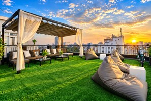 Una terraza en la azotea con zona de estar cubierta, césped artificial verde y vistas al atardecer.