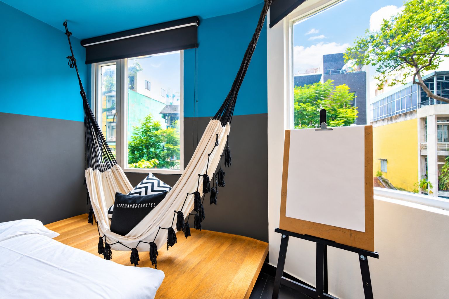 A room with a hammock, a bed, and a blank easel by a window with a city view.