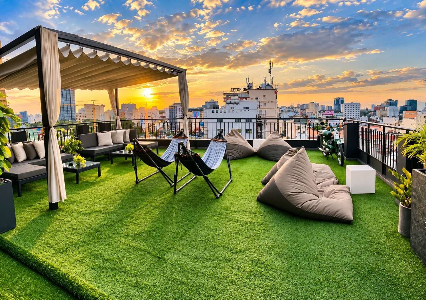 A rooftop terrace with green artificial grass, outdoor seating, hammocks, and a city skyline view at sunset.