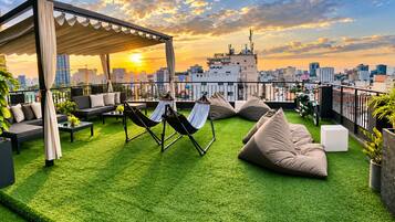 A rooftop terrace with green artificial grass, outdoor seating, hammocks, and a city skyline view at sunset.