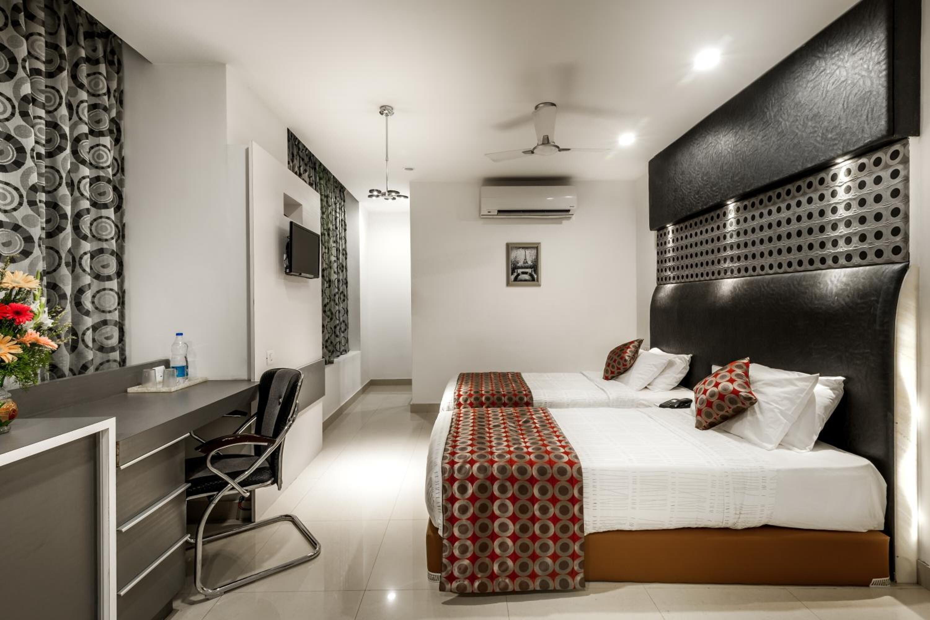 A modern hotel room with a desk, two beds, a ceiling fan, an air conditioning unit, and a decorative wall panel.