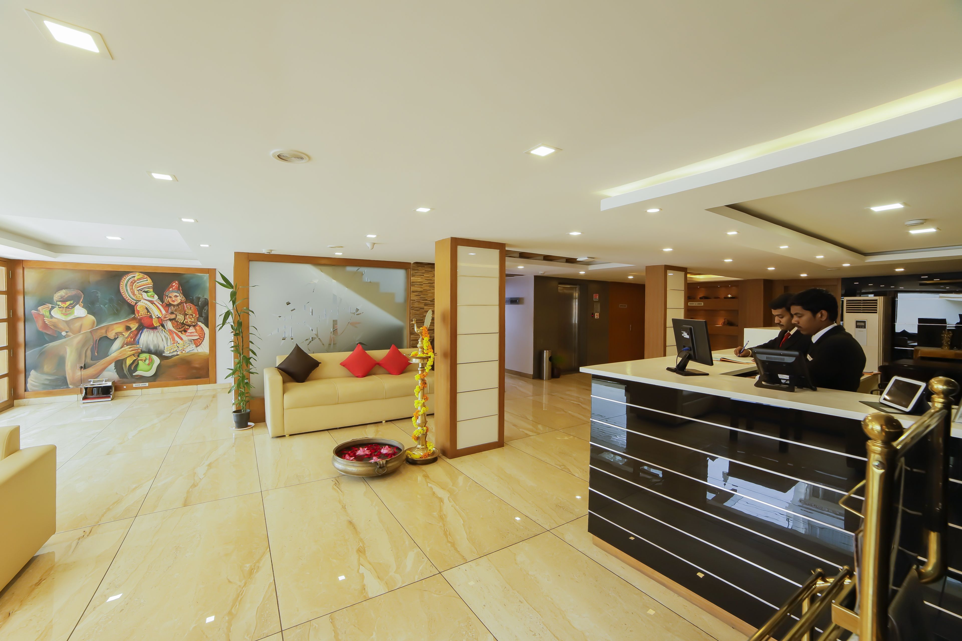 A hotel reception area with a reception desk, a sofa with red cushions, a decorative plant, and a mural on the wall.