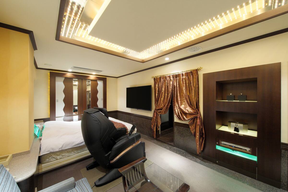 A hotel room with a bed, a massage chair, a flat-screen TV, a vanity with a mirror, and a wall-mounted shelf.