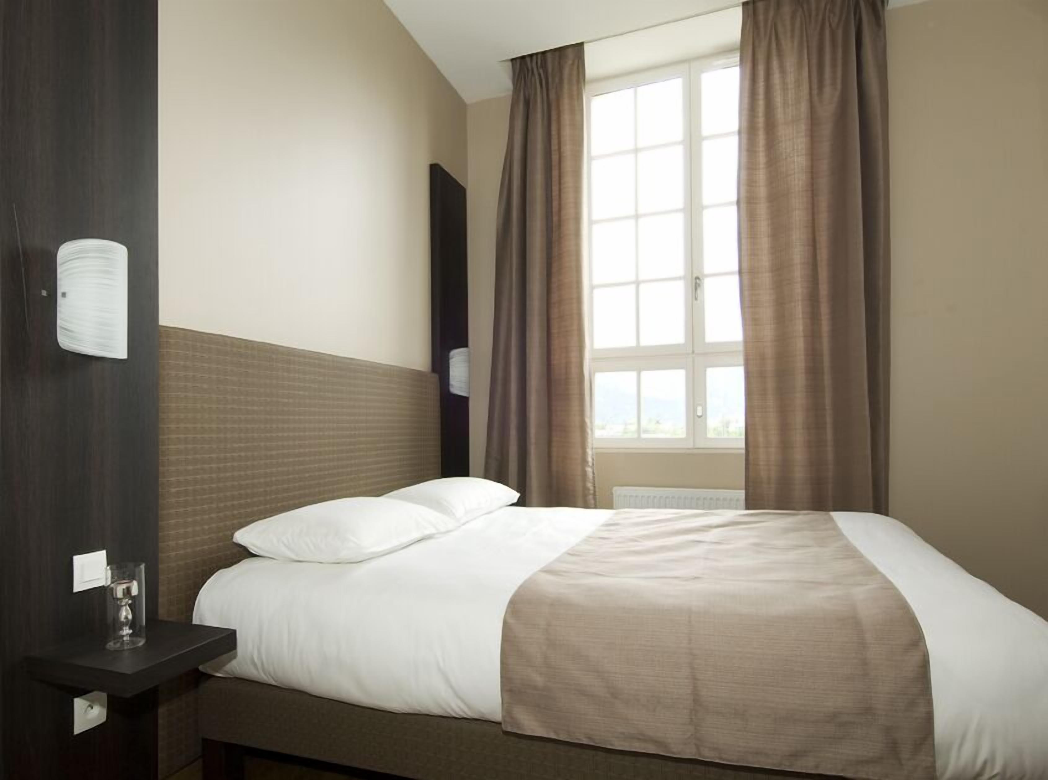 A hotel room with a large bed, a bedside table with a glass, and a window with curtains.