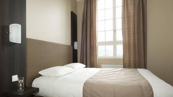A hotel room with a large bed, a bedside table with a glass, and a window with curtains.