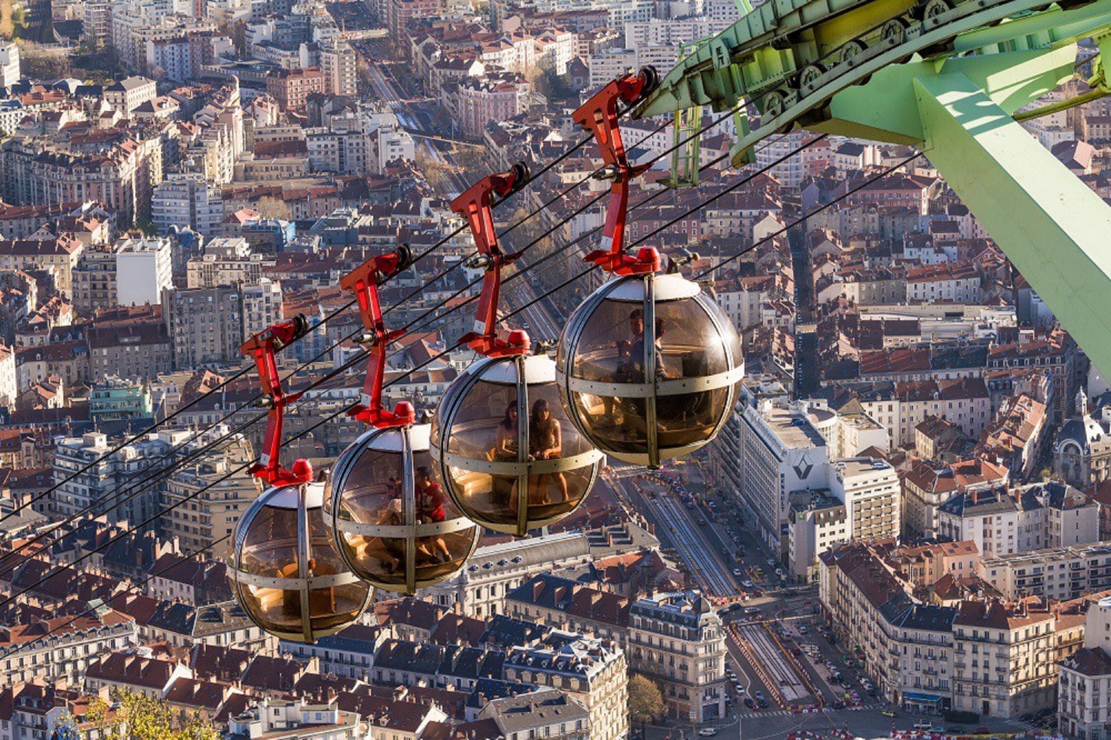 Sistema de teleférico com gôndolas esféricas suspensas por cabos vermelhos, proporcionando vista de uma cidade densamente construída.