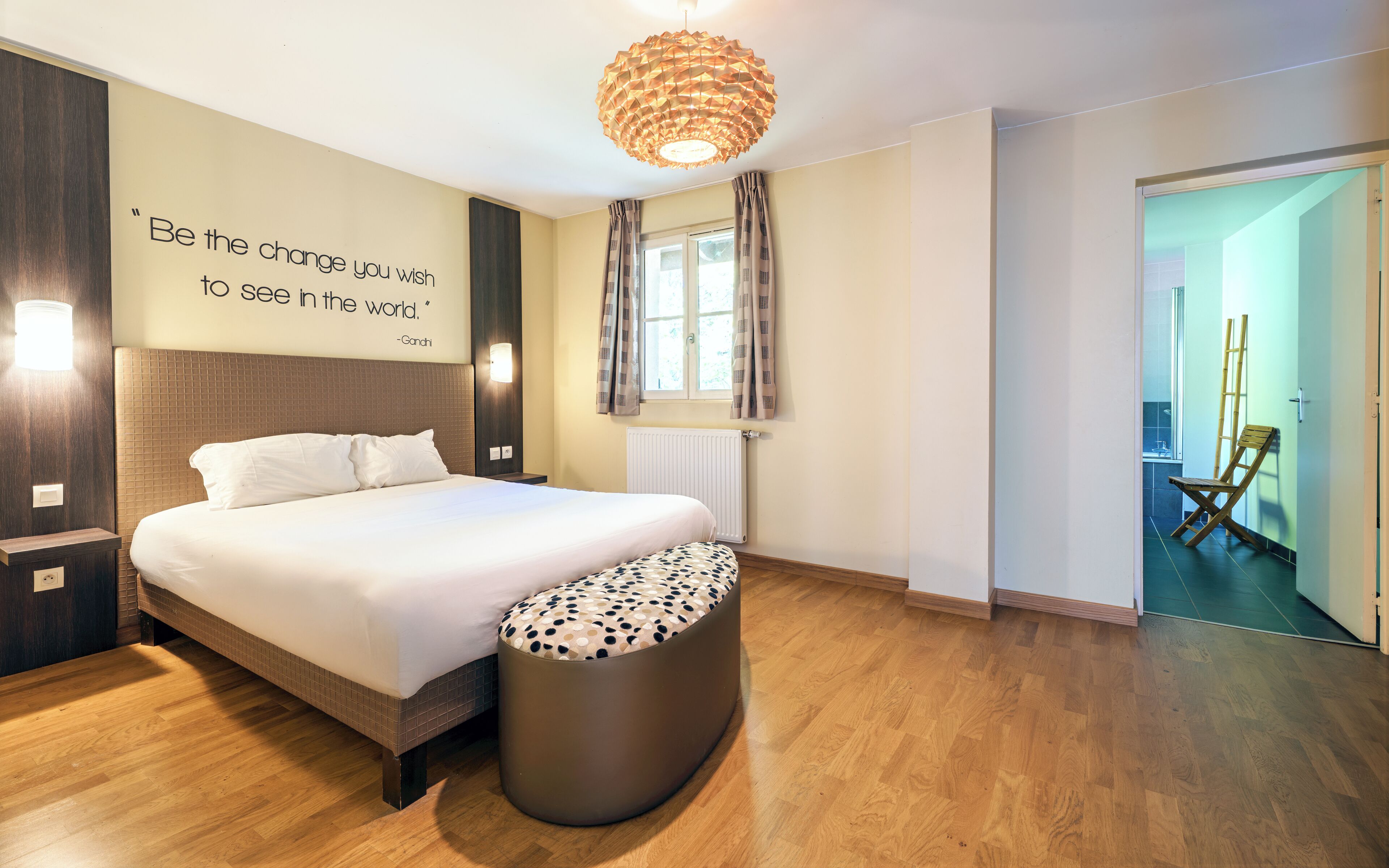 Moderni hotellihuone, jossa on suuri sänky, yöpöytä, seinään kiinnitetty valaisin, verhoilla varustettu ikkuna, seinässä oleva inspiroiva sitaatti ja puulattia.