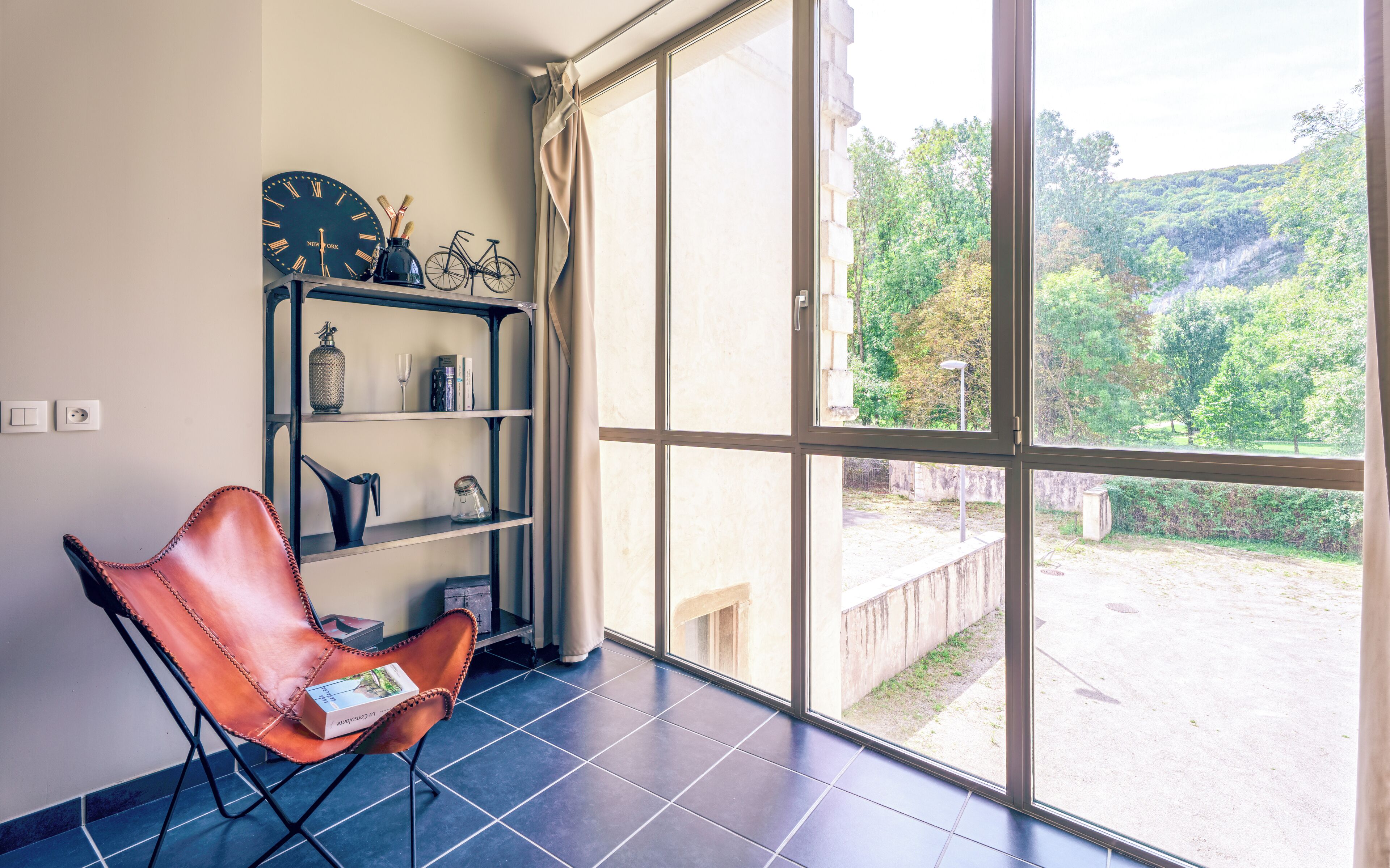 A room with a large window, a bookshelf, a leather chair, and a view of a green landscape.