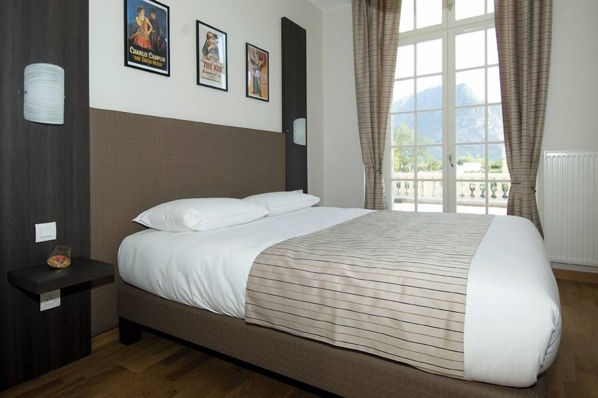 Quarto com uma cama grande, uma mesinha de cabeceira e uma janela com cortinas. Há pôsteres na parede e uma vista para as montanhas.