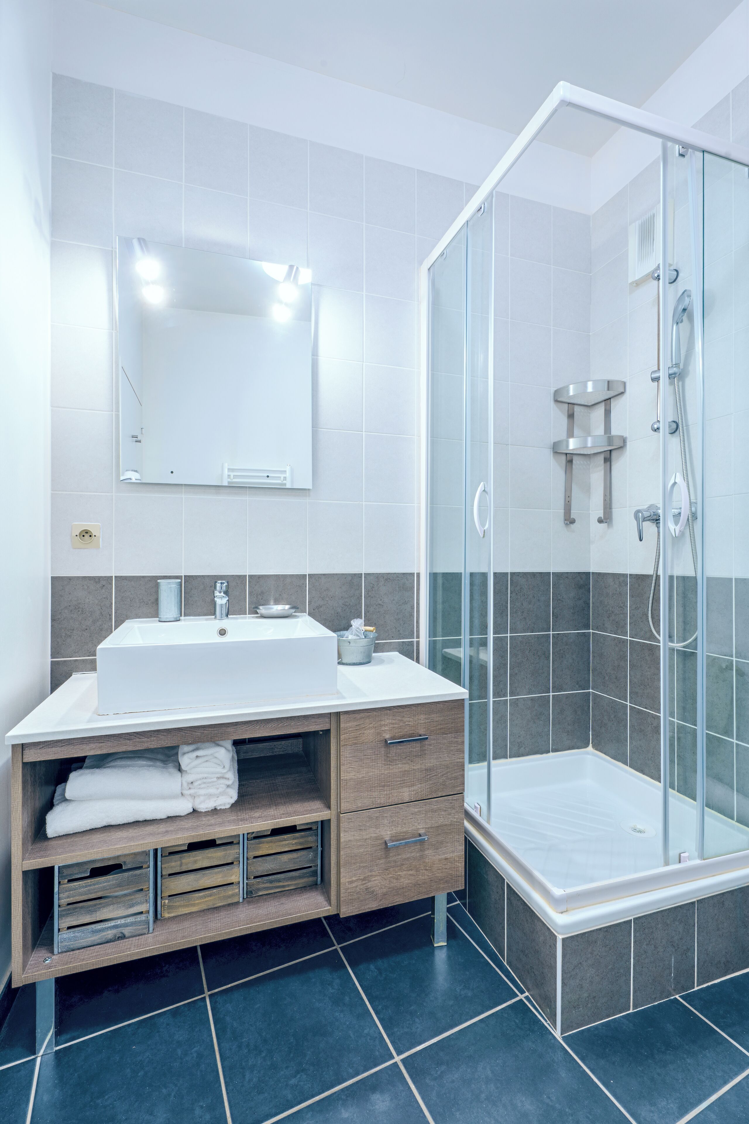 A modern bathroom with a glass-enclosed shower, a white sink with a mirror above, and a wooden vanity with drawers and shelves.
