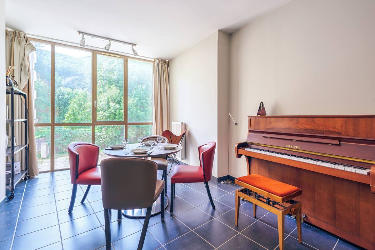 Quarto com piano, mesa de jantar com cadeiras e vista para o verde através de grandes janelas.
