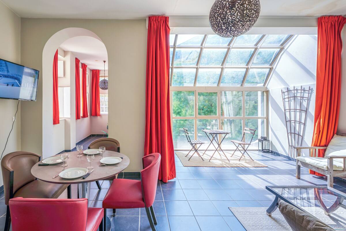 Uma área de jantar com mesa posta para quatro, cadeiras vermelhas, televisão na parede e vista através de grandes janelas.