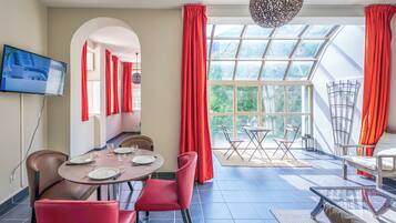 A dining area with a table set for four, red chairs, a TV mounted on the wall, and a view through large windows.