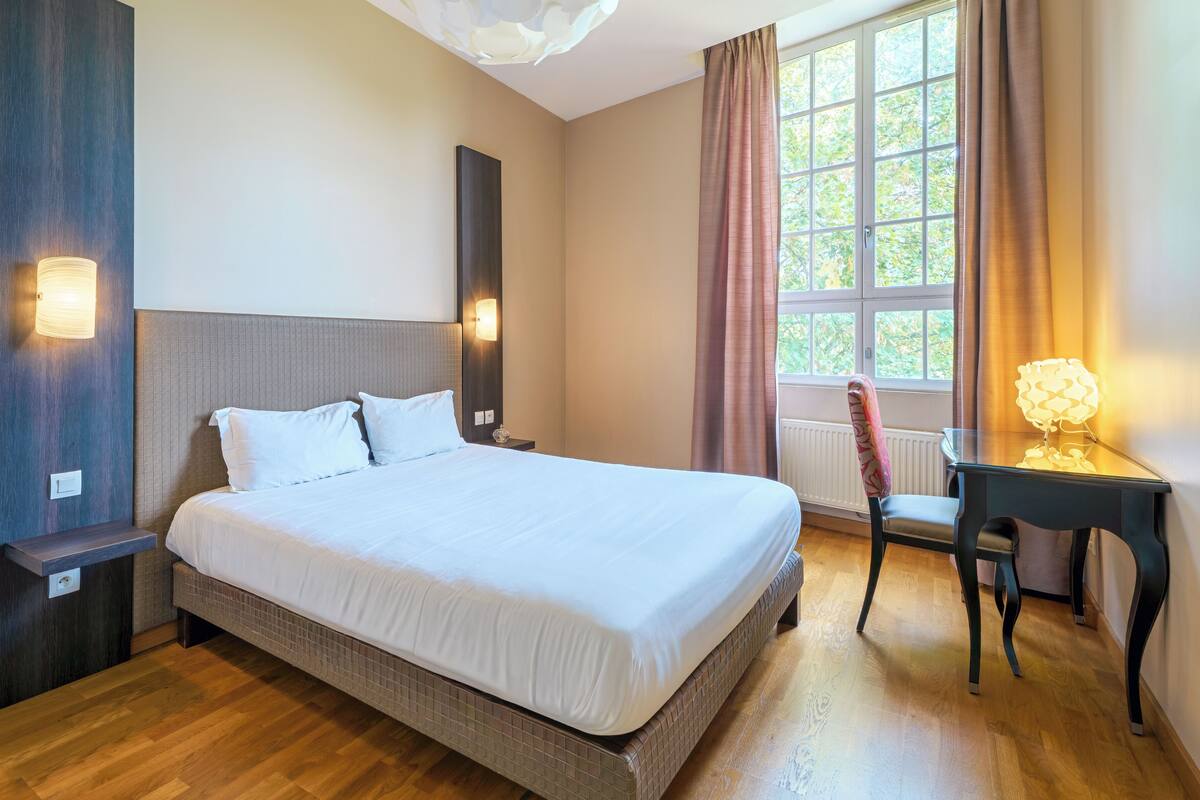 Quarto de hotel com cama, mesas de cabeceira, escrivaninha com luminária, cadeira e janela com cortinas.