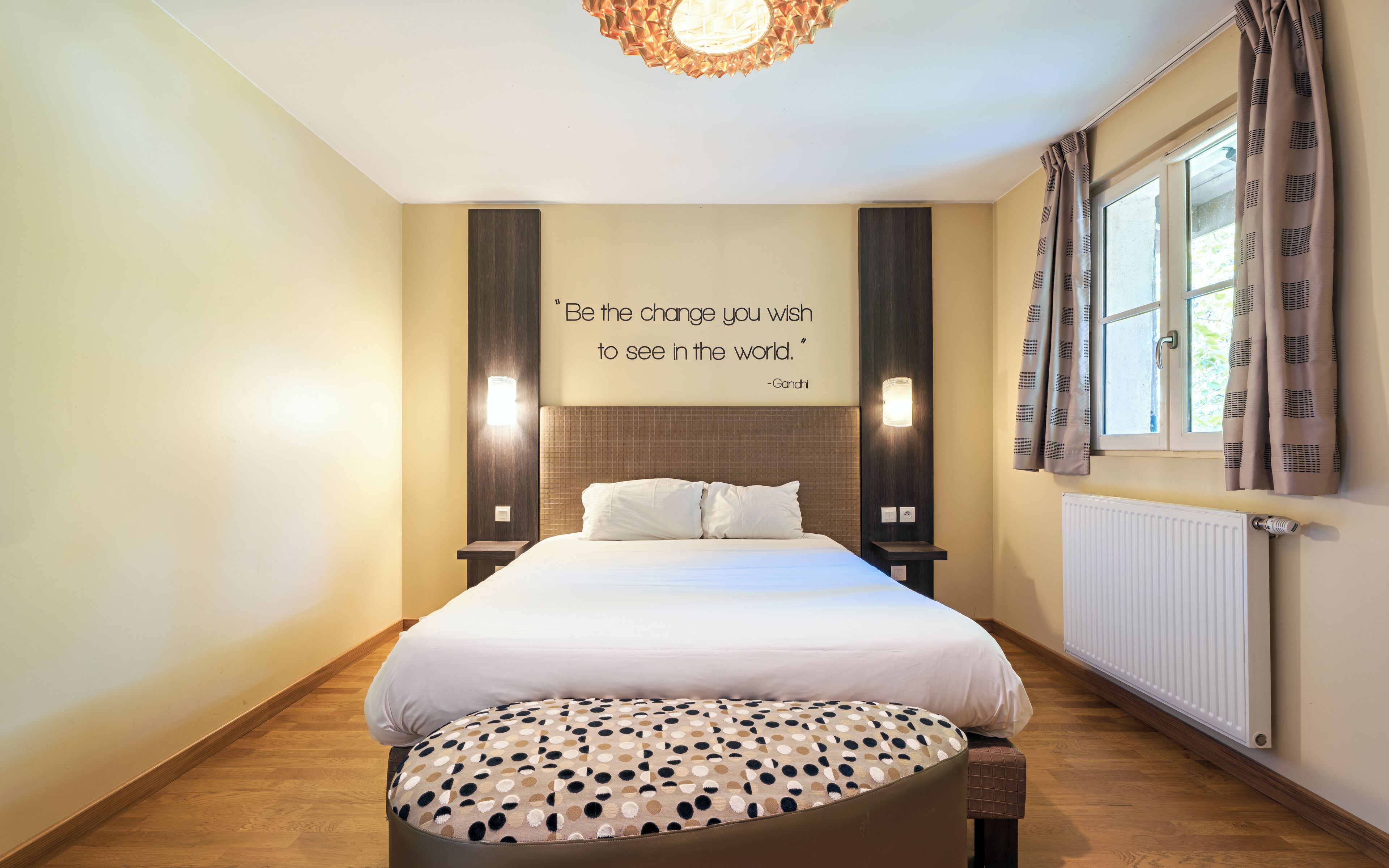 A hotel room with a bed, a quote on the wall, a window with curtains, and a radiator.
