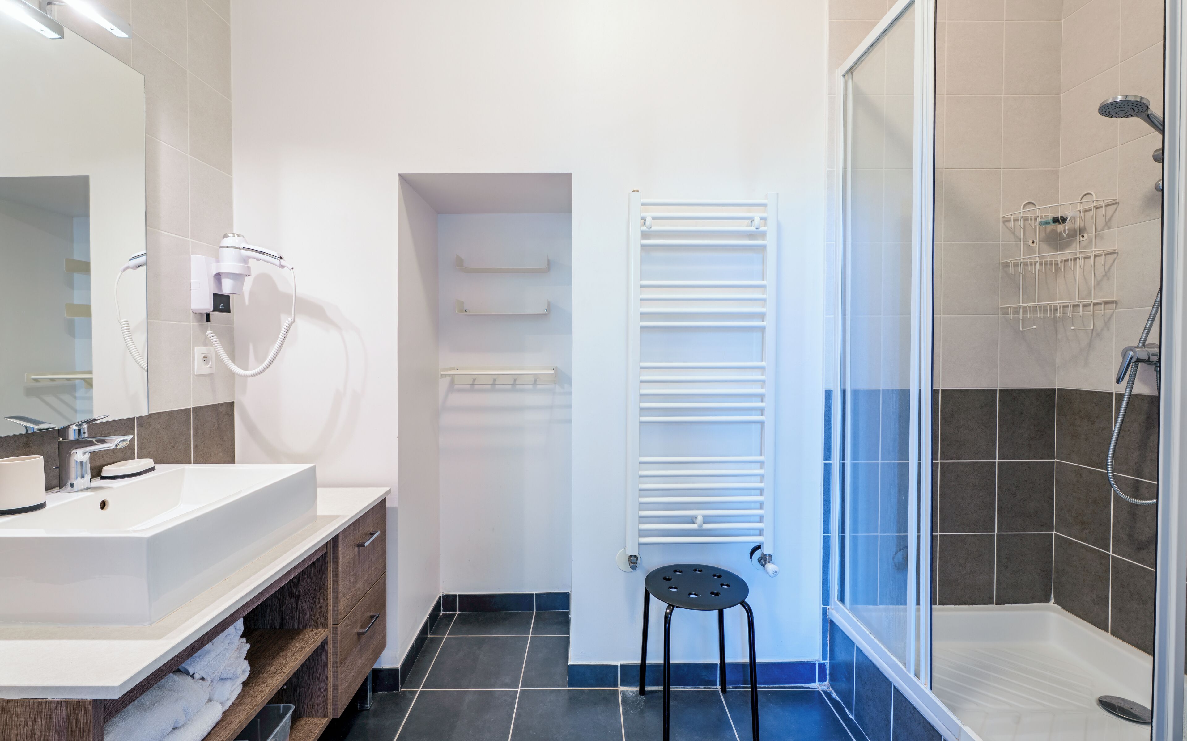 Een moderne badkamer met een wit wastafelmeubel, een haardroger, een handdoekrek, een douche met glazen deur en een zwarte kruk.