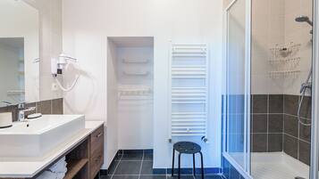 Een moderne badkamer met een wit wastafelmeubel, een haardroger, een handdoekrek, een douche met glazen deur en een zwarte kruk.